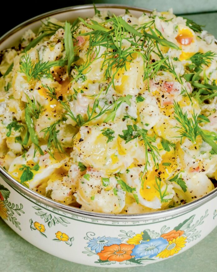 Potato salad, garnished with green herbs, sits in a casserole dish with blue, orange, and yellow flowers on it.