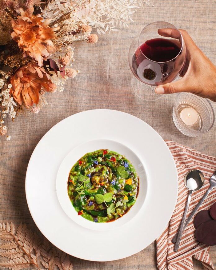 Escargot Risotto, a green looking dish, sits in a white bowl in the center of the photo. A white hand with a glass of red wine comes in from the right.