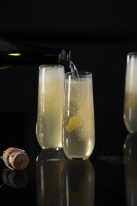 Two classic French 75 sit in champagne flutes against a black background with lemon peels inside the glass. A bottle of champagne pour into the glass to the right of the frame.