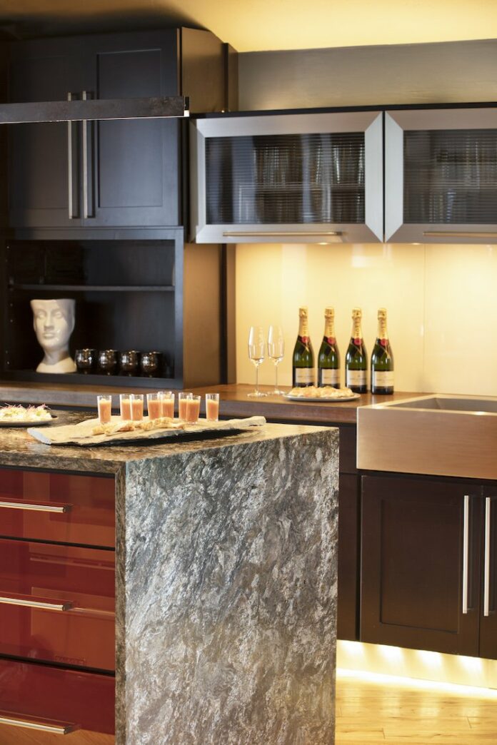 The interior of a kitchen with a granite waterfall island counter, and pink/orange looking drinks sitting on top. Behind the island is another countertop with four bottles of champagne.