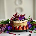 Strawberries, berries, and flowers adorn a ginger cake with the sides non-iced, blue, pink, and purples color the cake.