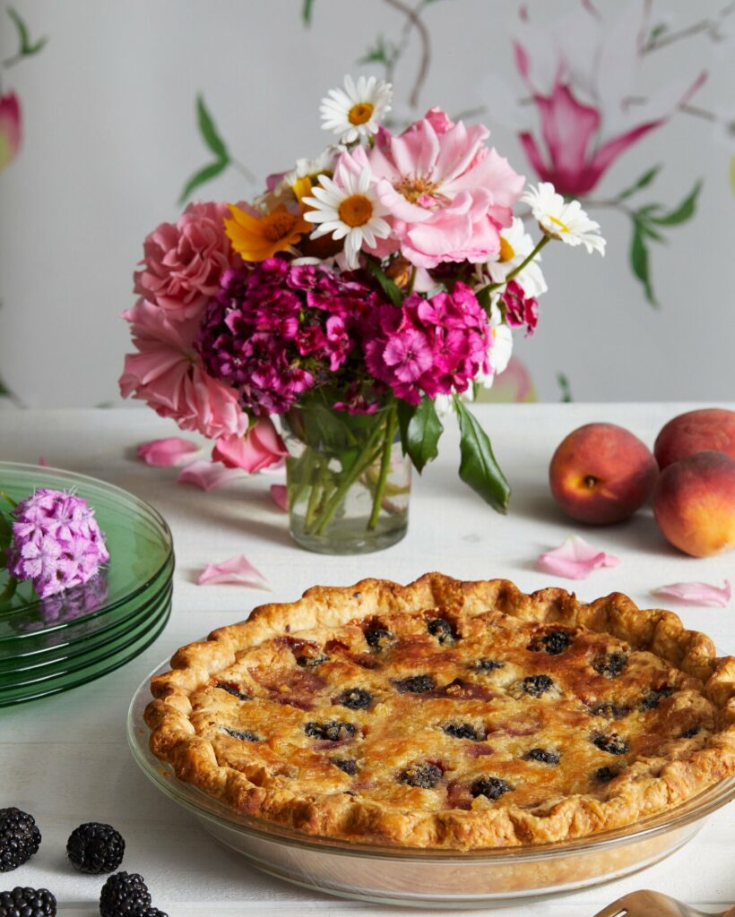 Gingered Peach and Blackberry Pie served in a glass plate with different-colored flowers in a glass on the side