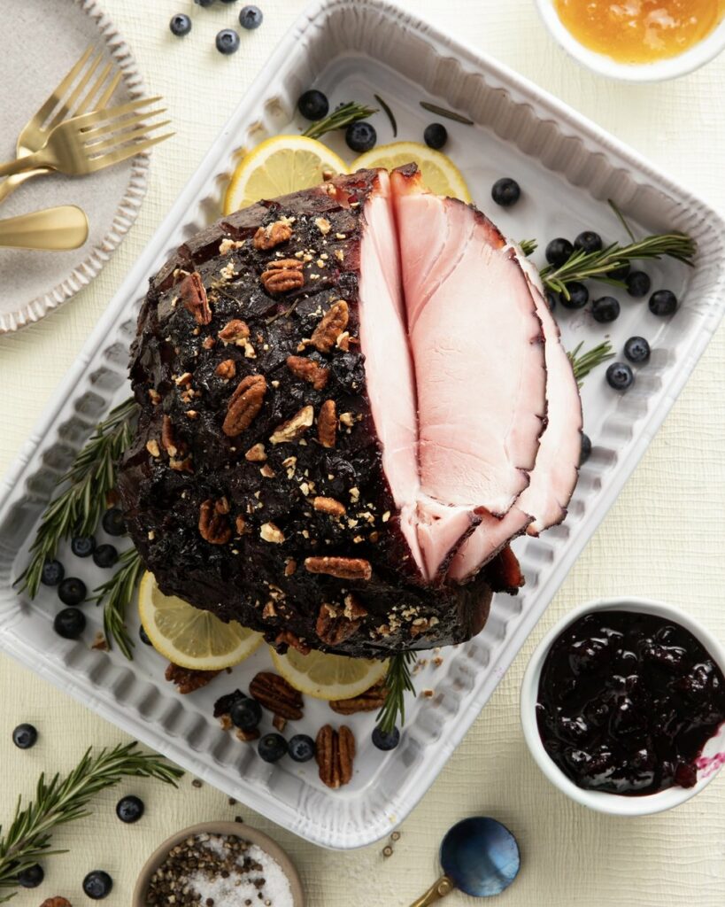 An aerial view of a cooked ham with blueberry yuzu glaze sitting in a white roasting tray with blueberries, thyme, and lemon garnishing the meat.