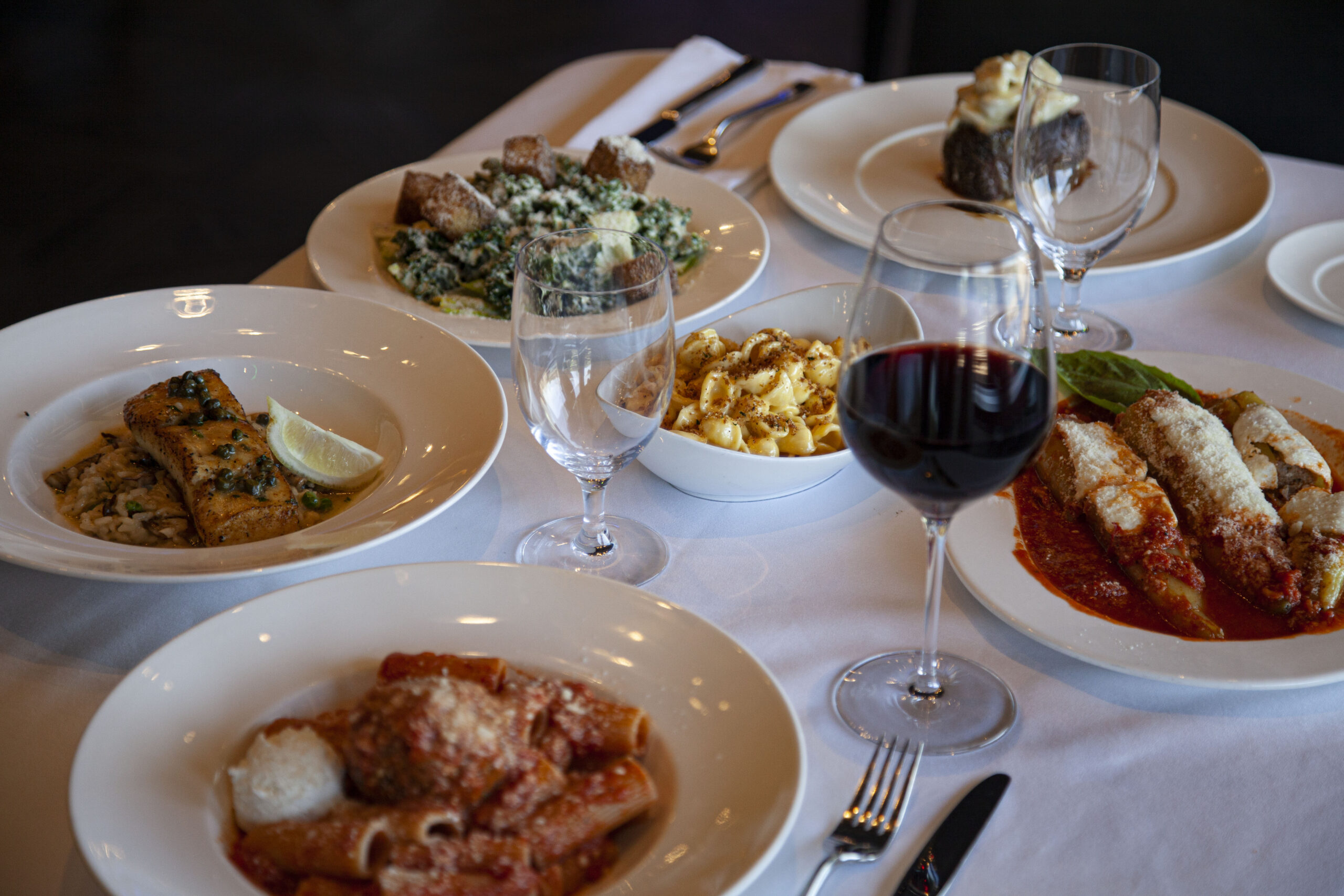 6 white plates of food on a white tablecloth with a glass of red wine and an empty wine glass