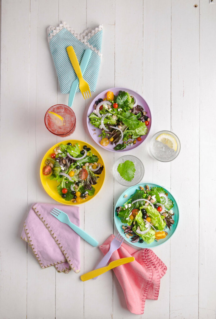 Three colorful bowls of kid-friendly summer salad with pink, purple, and blue napkins on the side.