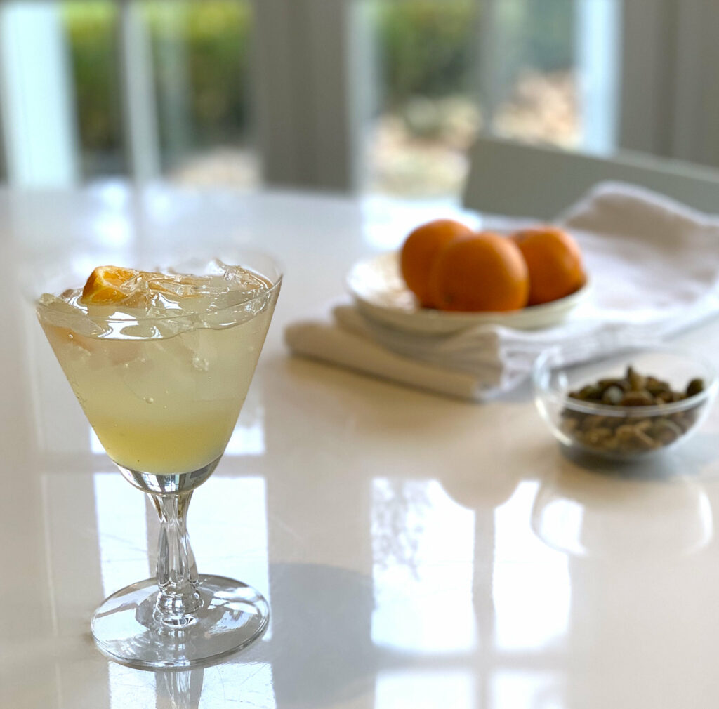 A lemon ginger shrub cocktail sits in a triangle glass, the liquid is light yellow in color. Behind the drink are three oranges on a cloth napkin.