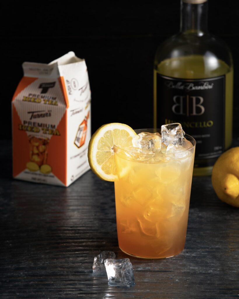 a cocktail garnished with a lemon slice on a dark surface with a carton of Turner's Premium Iced Tea behind it and a bottle of Limoncello.