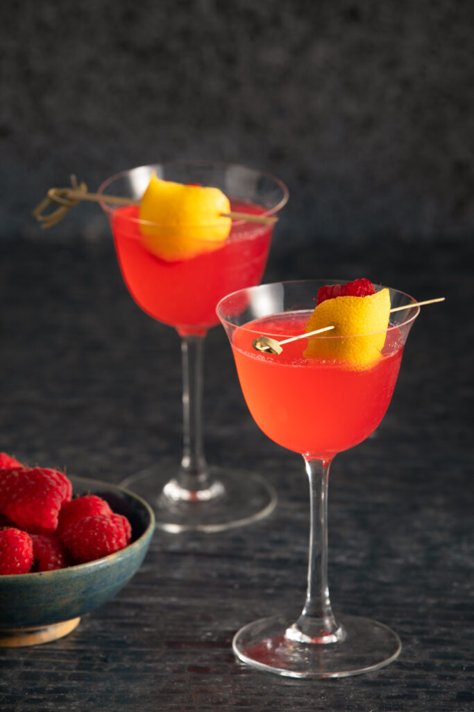 Moon Rising - Killers of the Flower Moon Cocktail. Two sit in martini glasses, garnished with a lemon peel. Bright red in color