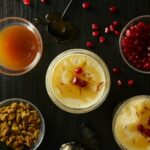 Golden Saffron Panna Cotta in small glass bowls with 3 additional bowls of nuts, caramel colored sauce, and pomegranate seeds.