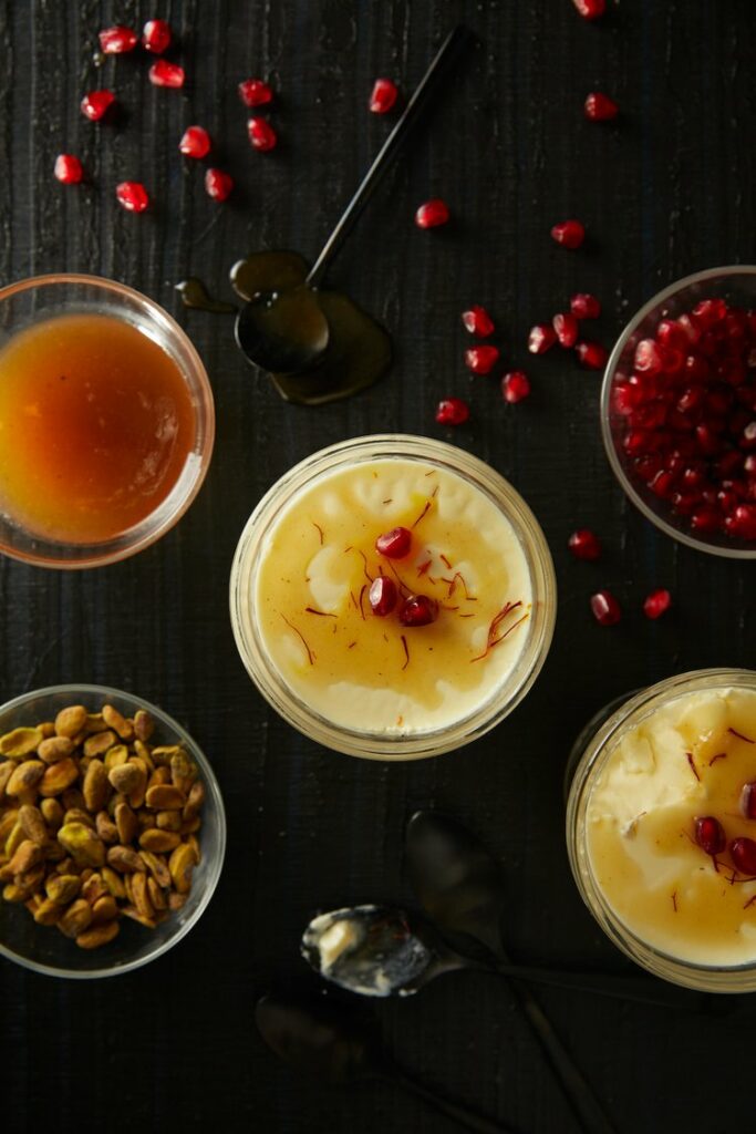 Golden Saffron Panna Cotta in small glass bowls with 3 additional bowls of nuts, caramel colored sauce, and pomegranate seeds.