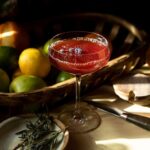 Sangre Sunset cocktail sits red in a coupe glass with a toothpick of blueberries floating on top.