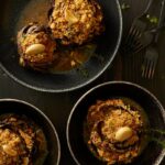Three stuffed artichokes in black bowls, with a garlic clove garnish and a sauce in the bottom of the bowl.