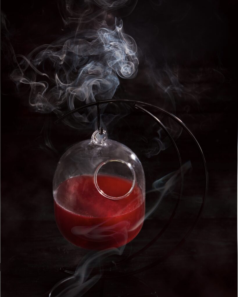 a deep red cocktail in a hanging glass with a small bird hole type opening with a dark background and smoke
