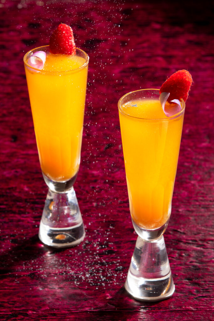 2 tall narrow orange cocktails with a pick sugar petal and raspberry garnish with a deep red background, Taylor Swift cocktails