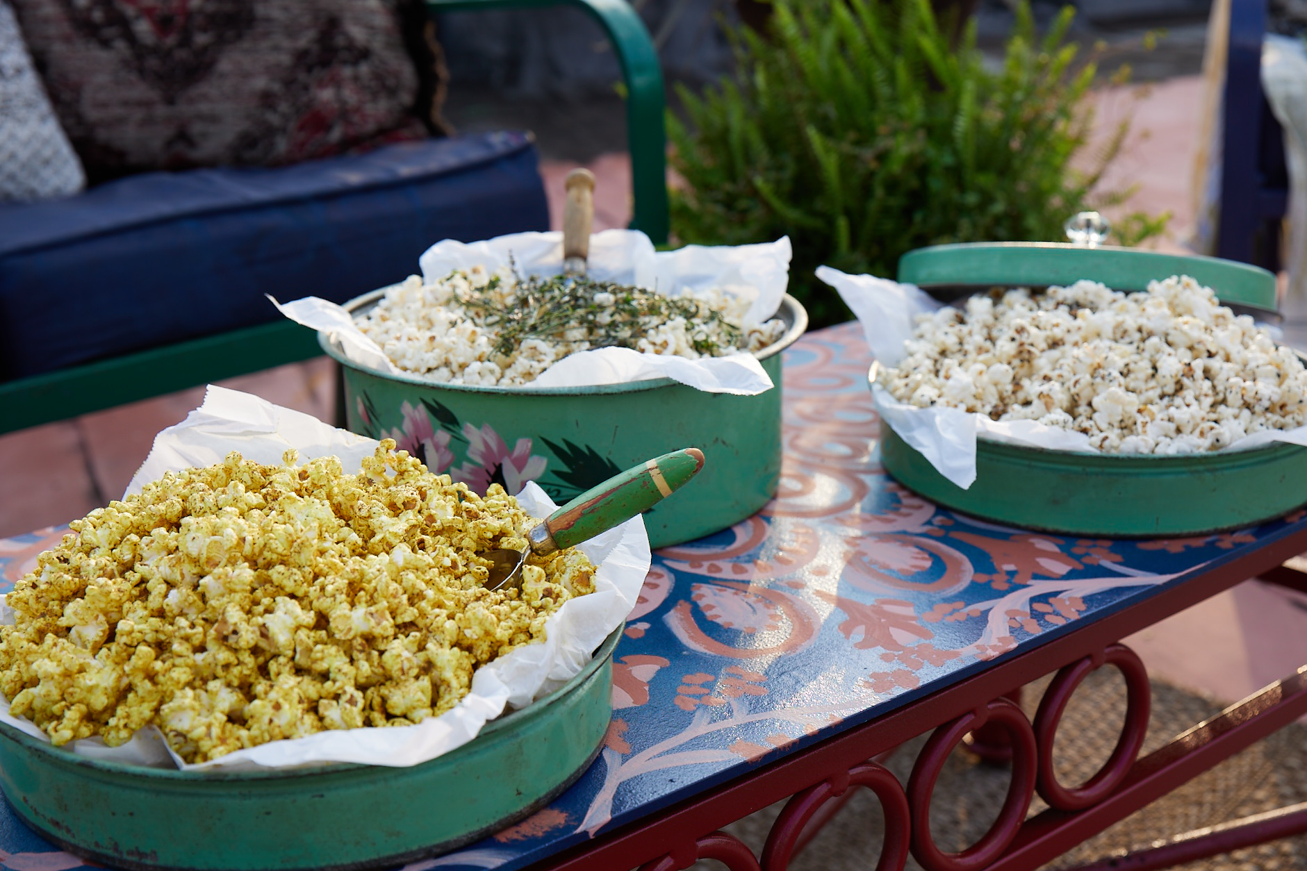 3 green antique metal bowls lined with white paper and filled with 3 different types of popcorn all sitting on a coffee table outside