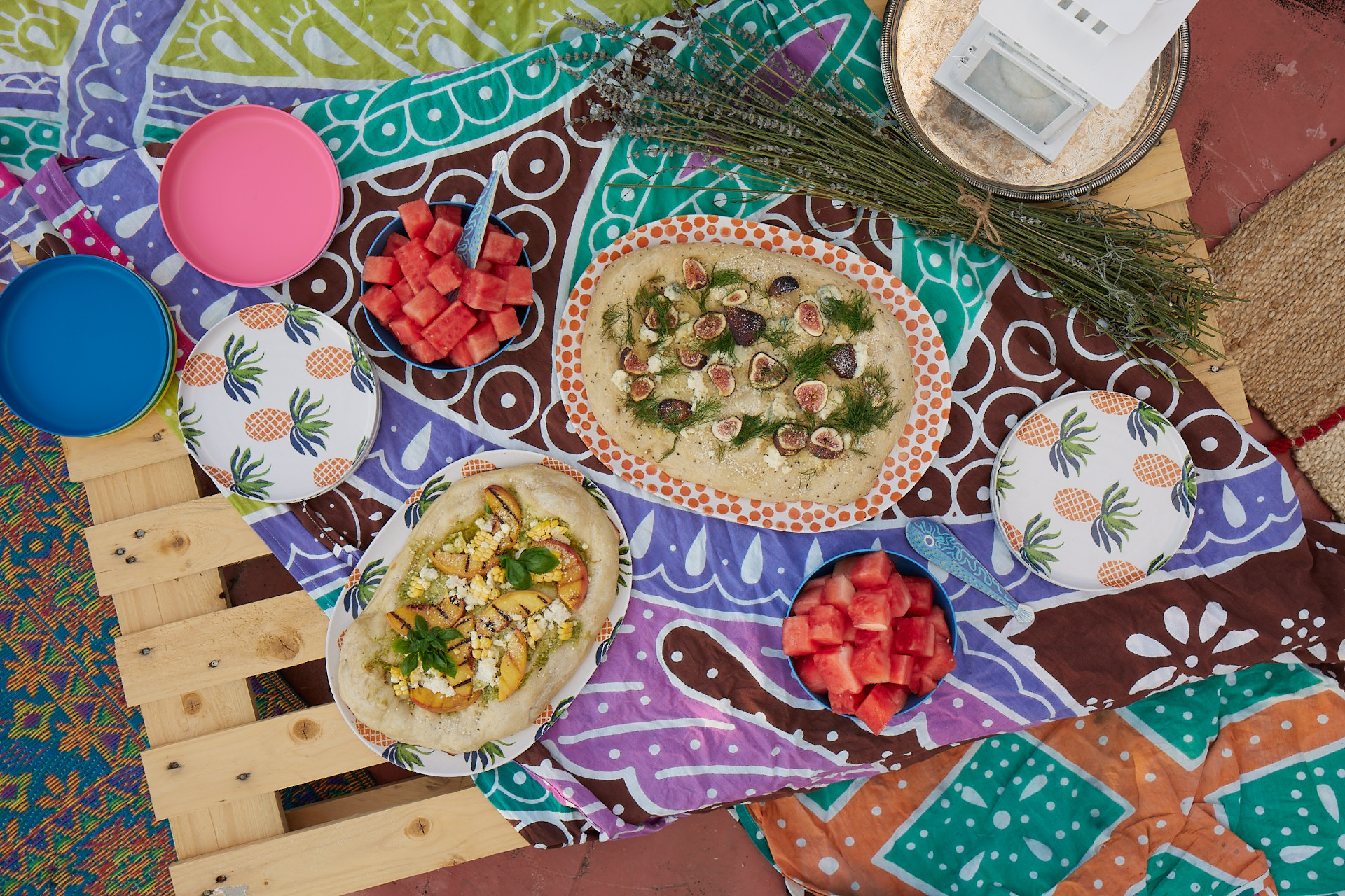 a fig flatbread and a corn and peach flatbread, both on colorful plates, a stack of pineapple printed plates on each side of the photo with two more plate stacks, one pink and one blue. Everything is sitting on a very colorful printed piece of fabric that is on a wooden pallet