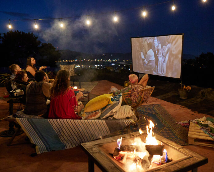 a few people sitting on furniture on a rooftop watching a movie on a screen with lights strung overhead and a small tabletop fire to the side