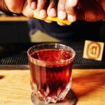 Unsung Heroes cocktail in a rocks glass, brown in color. A hand is positioned over the top, squeezing lemon into the drink.