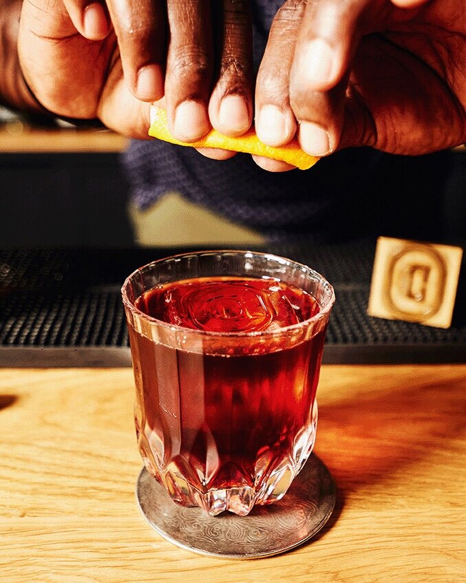 Unsung Heroes cocktail in a rocks glass, brown in color. A hand is positioned over the top, squeezing lemon into the drink.