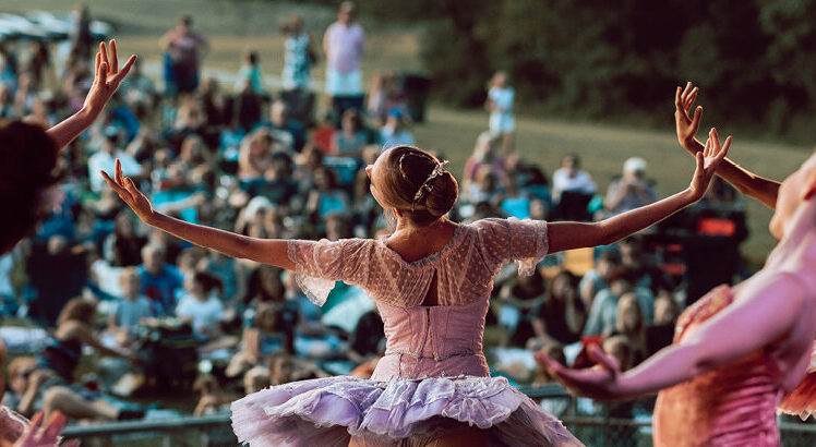 A ballerina with blond hair pulled in a tight buns leans back with her arms extended in front of an outdoor crowd