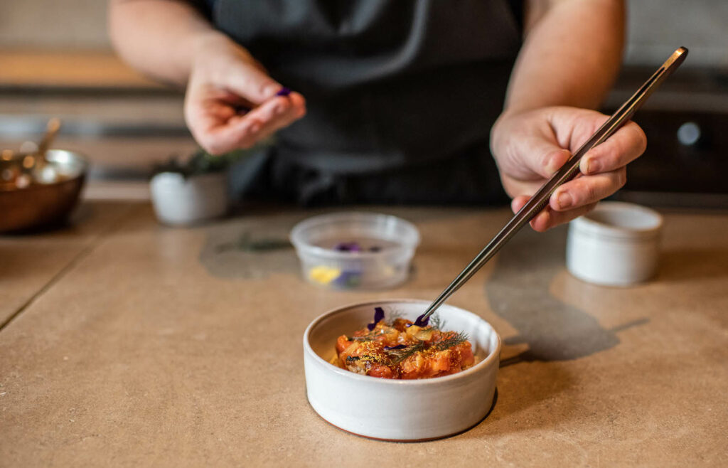 The bottom half of a person stands behind a counter and garnishes Crispy Brown Rice with Blue Crab, Smoke Tomato Vinaigrette and Fennel Purée with chopsticks. A delicious blue crab recipe