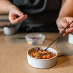 The bottom half of a person stands behind a counter and garnishes Crispy Brown Rice with Blue Crab, Smoke Tomato Vinaigrette and Fennel Purée with chopsticks. A delicious blue crab recipe