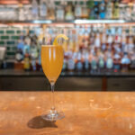 A cocktail brown-orange in color sits in a champagne flute on a bar. Blackberry Farm's Barn 75 Cocktail Recipe