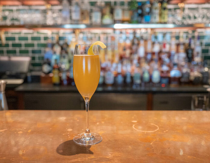 A cocktail brown-orange in color sits in a champagne flute on a bar. Blackberry Farm's Barn 75 Cocktail Recipe