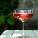 A cocktail red in color in a rounded martini glass sits on a white wooden slated table outdoors. Blackberry Farm's Blackberry Bumpkin Cocktail