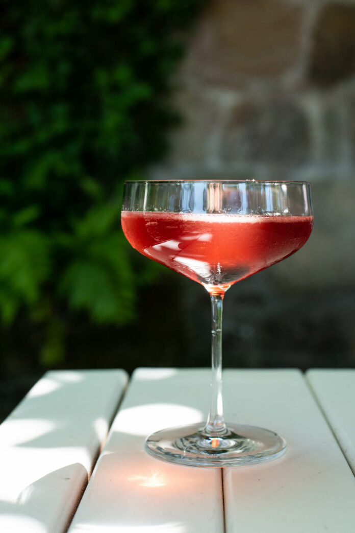 A cocktail red in color in a rounded martini glass sits on a white wooden slated table outdoors. Blackberry Farm's Blackberry Bumpkin Cocktail