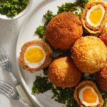 Chorizo Scotch Eggs with Chimichurri sits on a white plate surrounded by greenery.