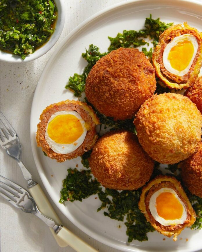 Chorizo Scotch Eggs with Chimichurri sits on a white plate surrounded by greenery.