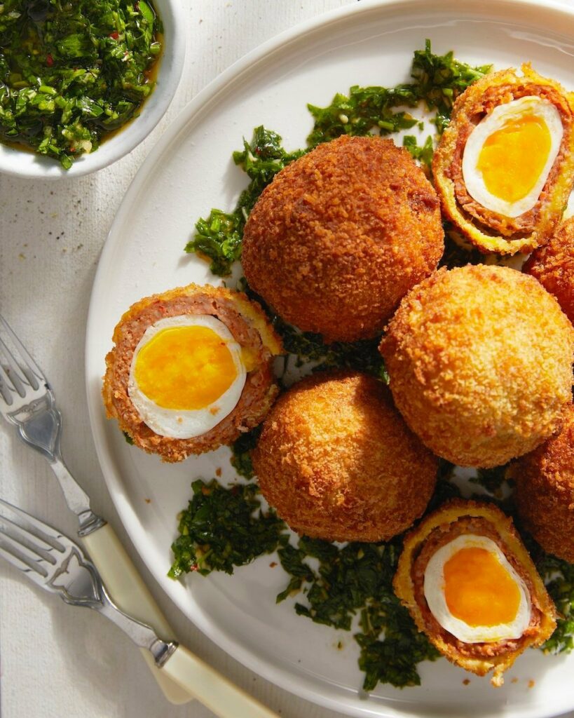 Chorizo Scotch Eggs with Chimichurri sits on a white plate surrounded by greenery.