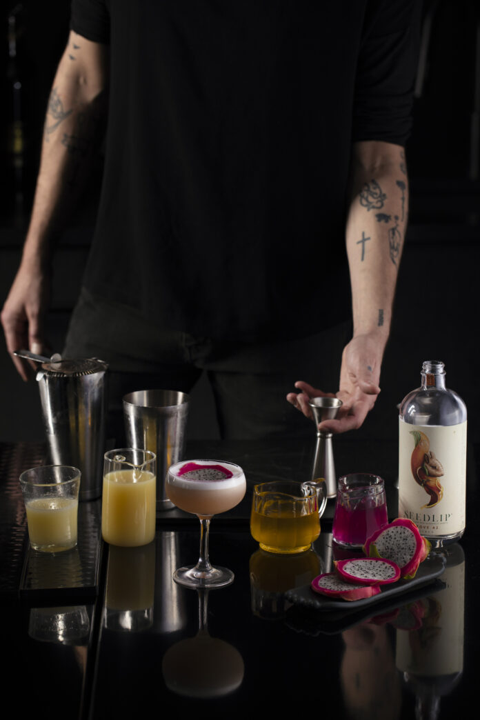 Mocktail Recipes - TABLE Magazine A variety of liquids, orange and pink in color, sit on a black table. The torso of a white mans leans forward and holds a jigger.