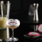 A martini mocktail light orange-y/pink in color sits in a martini glass next to sliced dragonfruit.