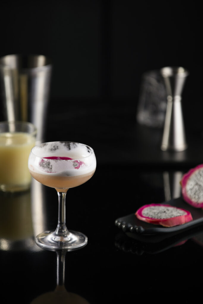 A martini mocktail light orange-y/pink in color sits in a martini glass next to sliced dragonfruit.