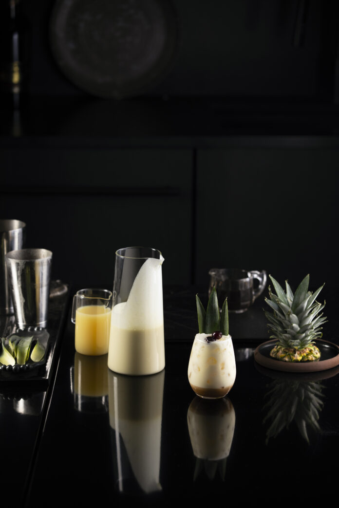 A piña colada mocktail, white in color, sits in a stemless wine glass next to a glass pitcher of the mixed drink.