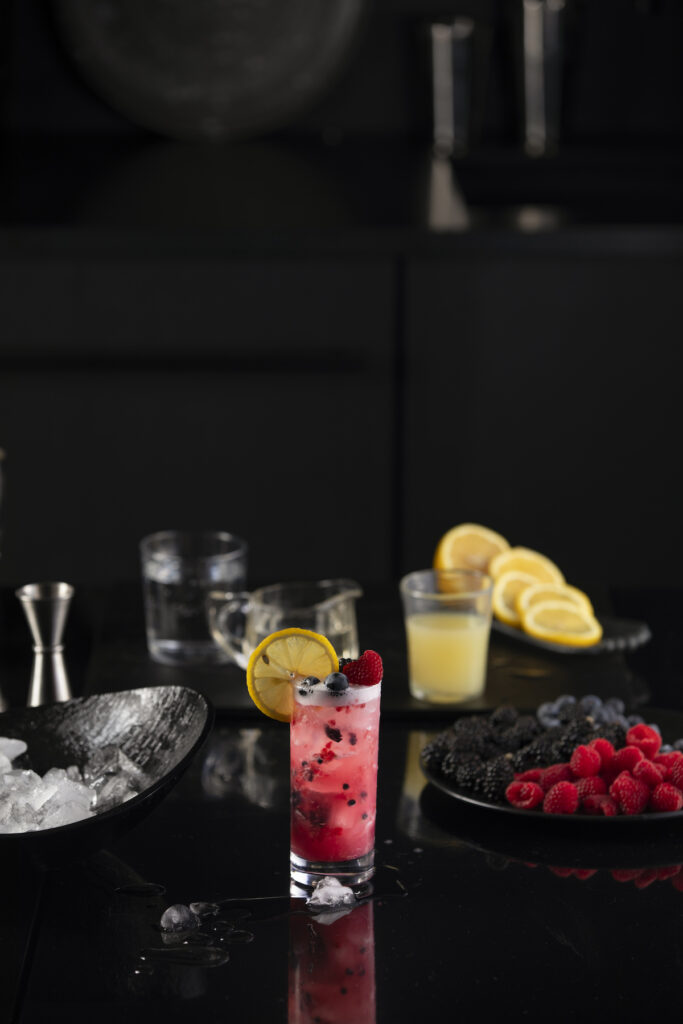A beverage red in color, sits in a glass filled with berries and garnished with lemon.