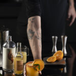 A white man's arm and hand holds a Spicy Orange NA-rgarita (a Margarita Mocktail) on a black filled with mocktail mixing supplies.