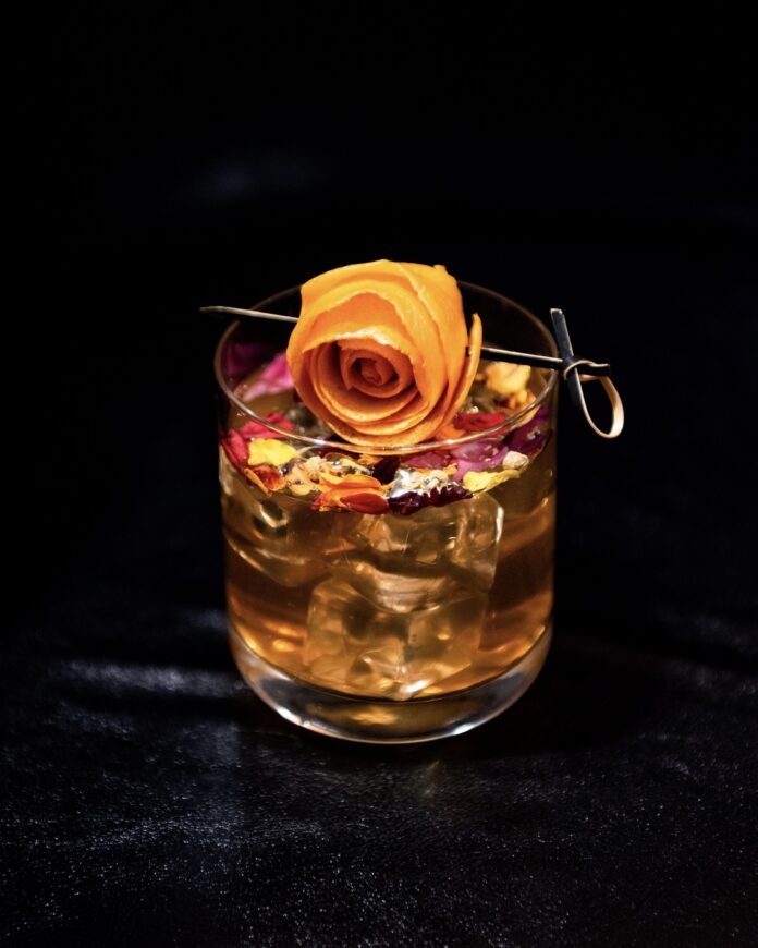 a bourbon drink in a rocks glass with an orange peel garnish that looks like a rose resting atop a glass with dred flower petals and a black background