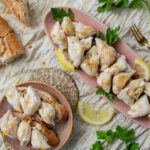 large lump crab pieced covered in brown butter and lemon zest on a small round pink plate and a long narrow pink tray with a broken crusty baguette, parsley, and lemon slices on a piece of light yellow spotted fabric