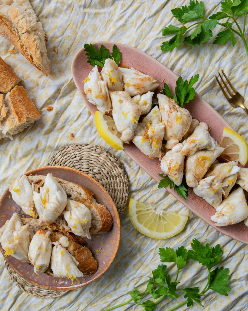 large lump crab pieced covered in brown butter and lemon zest on a small round pink plate and a long narrow pink tray with a broken crusty baguette, parsley, and lemon slices on a piece of light yellow spotted fabric