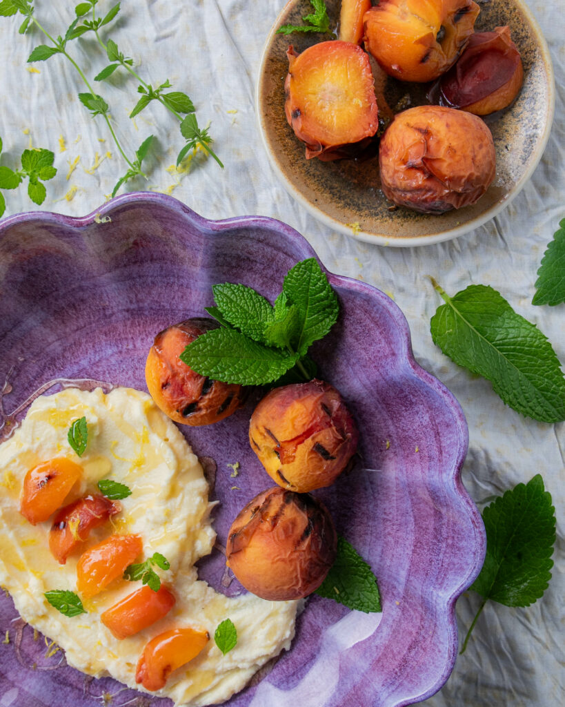 an arial view of a wide purple bowl with grilled peaches, whipped mascarpone and honey, garnished with fresh mint, and a smaller neutral colored bowl with grilled peaches