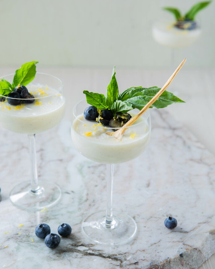 Lemon Basil Posset a 3/4 look in photo of 3 Lemon basil possets in clear coupe glasses garnished with blueberries and basil on a marble base with a white background