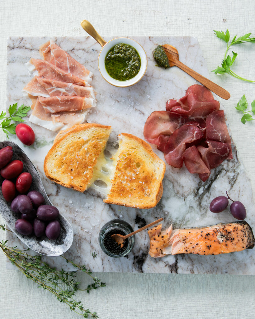 a grilled cheese in the middle of a marble charcuterie board with red and purple olives and a variety of cured meats