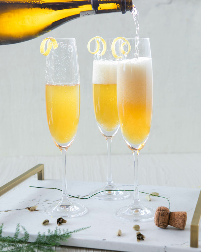 3 champagne flutes being filled with Cognac and Cardamom French 75 cocktails on a Cambria surface with a white background