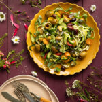 An aerial shot of a colorful summer salad on a yellow plate sitting on wooden slates purple in color and surrounded by flowers and herbs. Summer Salad Recipe