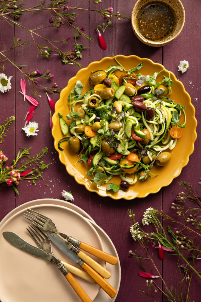 An aerial shot of a colorful summer salad on a yellow plate sitting on wooden slates purple in color and surrounded by flowers and herbs. Summer Salad Recipe