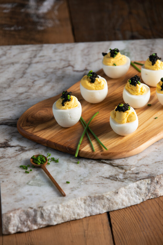 a wooden board on a piece of white and gray marble on a wooden table with six deviled eggs topped with caviar and garnished with chives