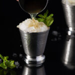 A white hand pours a mint julep into a silver julep cup.